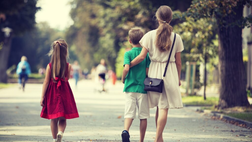 Back view of young blond long-haired woman walking with two children, small daughter and son along sunny park alley on blurred people silhouette and green trees bokeh background on warm summer day.